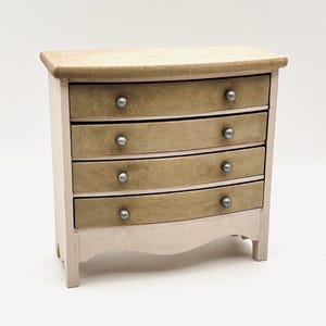 May include: Miniature wooden dresser with four drawers. The drawers are a light brown color with silver knobs. The frame is painted a pale pink color. The top of the dresser is a natural wood color.