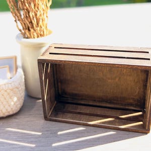 May include: A small, rectangular wooden crate with a dark brown finish. The crate has three horizontal slots on the top and is open on one side. A white ceramic vase with dried stems and a woven basket are also in the image.