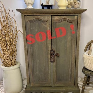 May include: A small, weathered-look wooden cabinet with two doors, a decorative handle, and the word "SOLD!" in red. The cabinet is on a table with a lamp, a framed picture, and other decorative items.