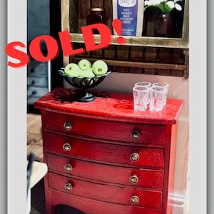 May include: Miniature red dresser with four drawers and brass knobs. A shelf above holds white teacups, coffee, and decorative items. A bowl of green apples and small glasses sit on top. Text reads "SOLD" and "Do Not Buy - Sample Work!"