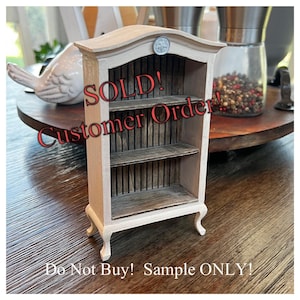 May include: A miniature wooden bookshelf with two shelves and a decorative top. The bookshelf is painted in a light beige color with a distressed finish. The text "SOLD! Customer Order! Do Not Buy! Sample ONLY!" is written on the front.
