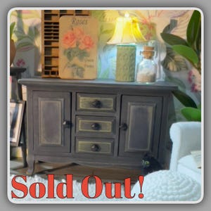 May include: Miniature gray cabinet with three drawers and two doors, accented with gold trim. A small lamp, a decorative bottle, and a sign that says "Roses" sit on top. The text "Sold Out!" is displayed at the bottom.