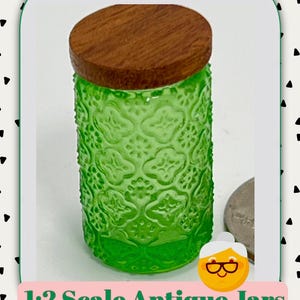 May include: A green glass jar with a wooden lid. The jar has a textured floral pattern and is labeled "1:2 Scale Antique Jars". The jar is cylindrical and has a round wooden lid.