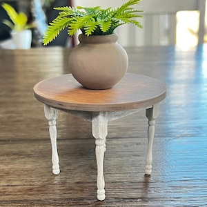 May include: A miniature round table with a brown tabletop and white legs. A small, tan vase holds green leafy plants. The table is a decorative piece, likely for a dollhouse or similar display.