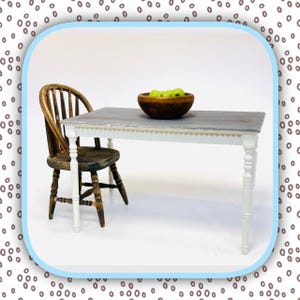 May include: A miniature dining set featuring a gray-topped table with white legs and a wooden chair. A wooden bowl filled with green apples sits on the table. The table is approximately 12 inches long.