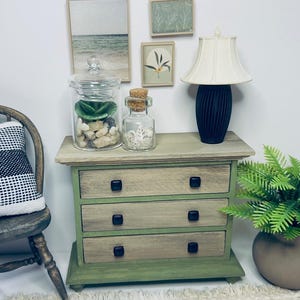 May include: Miniature wooden dresser with three drawers, painted in shades of green and natural wood. The dresser is adorned with black square knobs. A glass jar with a lid, filled with rocks and a succulent, sits on top, along with a small lamp.