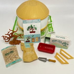 May include: A bonsai kit with a yellow and white container, a small red tray, and a miniature shovel. The kit includes a bag of soil, a packet of seeds, and a decorative bonsai tree. The packaging includes the text "JUNIPER BONSAI".