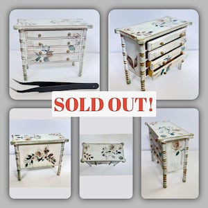 May include: Miniature white painted chest of drawers with four drawers, floral accents, and gold-colored hardware. The chest has a distressed finish and bamboo-style legs. The text "SOLD OUT!" is displayed in red.