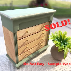 May include: A small wooden chest of drawers with four drawers, painted in a two-tone color scheme of light green and natural wood. The chest has brass knobs and is labeled "SOLD!" and "Do Not Buy - Sample Work!"