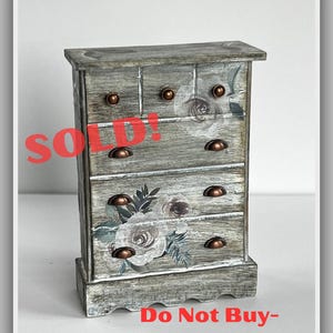 May include: A small, gray wooden cabinet with five drawers and copper-colored hardware. The cabinet is decorated with floral decals and the word "SOLD!" is printed in red. The text "Do Not Buy - Sample Work!" is also visible.