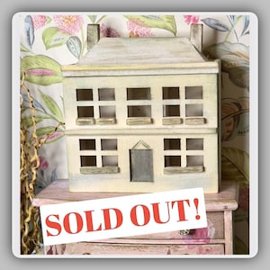 May include: A two-story, off-white wooden house with multiple square windows and a gray door. The roof and window frames have a weathered appearance. A sign reading "SOLD OUT!" is in front of the house. The house is placed on a pink and floral patterned surface.