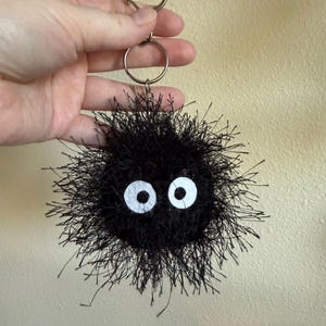 May include: A black fuzzy circular keychain with long, spiky yarn fibers and two large, round white eyes with black pupils. The keychain is attached to a silver-toned metal ring and a heart-shaped clasp, creating a round, textured character design.