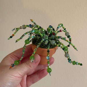 May include: A miniature beaded plant in a small terracotta pot. The plant features beaded stems and leaves in shades of green, with accents of blue, orange, and clear beads. The pot is filled with green moss.