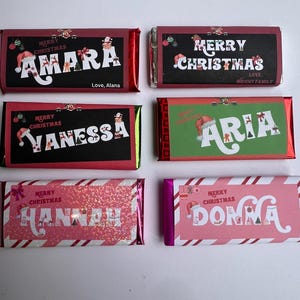 Op de afbeelding: Zes gepersonaliseerde chocolade repen verpakt in feestelijk rood-wit gestreept folie. Elke reep heeft een andere naam, waaronder Amara, Vanessa, Aria, Hannah en Donna. De repen zijn versierd met kerstthema illustraties en de tekst "Merry Christmas".