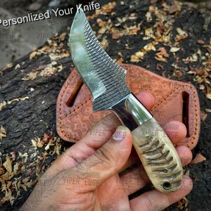 May include: A small, personalised knife with a file-like blade and a bone handle. The knife is held in a hand and is resting on a leather sheath. The sheath has a decorative pattern and the text "VIPER EDGE" is visible on the sheath.