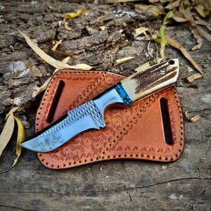Peut inclure: Un couteau à lame fixe avec une lame en acier poli dotée d'un dos quadrillé, une mitre à motif bleu et noir, et un manche en bois de cerf texturé, posé sur un étui en cuir beige avec des bords estampés décoratifs et des passants.