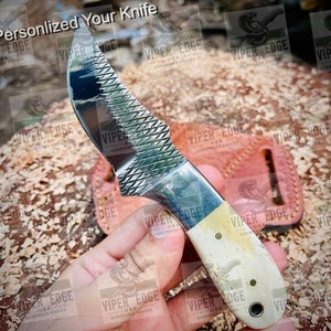 May include: A personalised knife with a white handle and a serrated blade. The blade has a textured pattern and the handle has a bone-like appearance. The knife is being held by a hand.