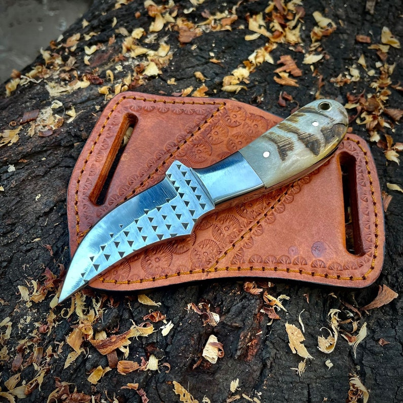 May include: A brown leather sheath with a decorative pattern holds a small, curved knife with a serrated blade and a bone handle.