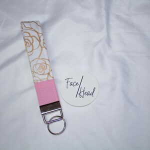 May include: A fabric wristlet keychain featuring a white strap with a gold rose outline pattern, a solid pink rectangular section, and a silver metal clasp with a circular ring. Next to it is a white circular tag with the text Face/Head.
