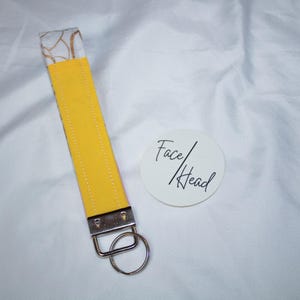 Peut inclure: Un porte-cl&eacute;s dragonne en tissu compos&eacute; d'une sangle jaune vif avec des coutures blanches, d'un fermoir en m&eacute;tal argent&eacute; et d'un anneau fendu circulaire. &Agrave; c&ocirc;t&eacute; se trouve un autocollant blanc rond avec le texte noir Face/Head imprim&eacute;.