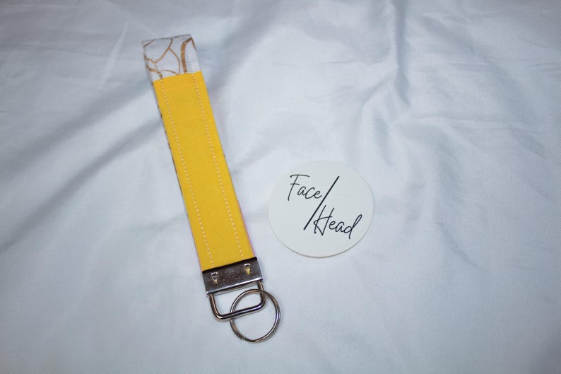 Peut inclure: Un porte-cl&eacute;s dragonne en tissu compos&eacute; d'une sangle jaune vif avec des coutures blanches, d'un fermoir en m&eacute;tal argent&eacute; et d'un anneau fendu circulaire. &Agrave; c&ocirc;t&eacute; se trouve un autocollant blanc rond avec le texte noir Face/Head imprim&eacute;.