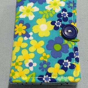 May include: A small, rectangular fabric case with a floral pattern in shades of blue, yellow, and white. The case is secured with a navy blue button and a green loop closure. The edges are neatly stitched.