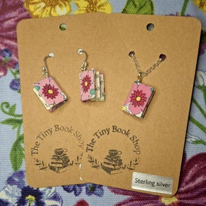 May include: Handmade book-shaped jewellery set, comprising earrings and a necklace. The miniature books have pink floral fabric covers with red and yellow flower designs. The earrings and necklace are displayed on a brown card with the text "The Tiny Book Shop".