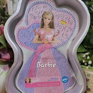 May include: A silver-coloured aluminium cake pan shaped like a doll in a gown. The insert features a blonde Barbie doll wearing a pink bodice and a purple and pink textured skirt. The text reads Barbie, Cake Pan, and Wilton, with instructions.