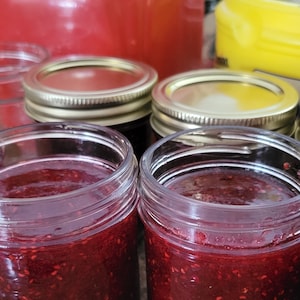 May include: Four glass jars filled with red raspberry jam. The jars have gold lids and are sitting on a brown surface.