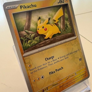 May include: A Pikachu Pokémon trading card, featuring a yellow Pikachu character with black stripes and red cheeks. The card has text including "Pikachu", "Charge", and "Pika Punch". The card is displayed in a clear plastic holder.