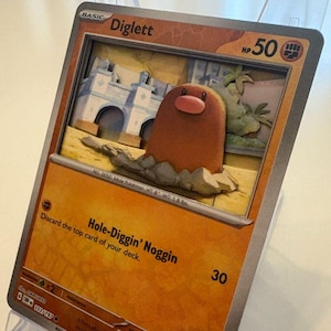 May include: A 3D artwork of a Diglett Pokémon card. The card features a brown Diglett emerging from the ground, with a light brown background and a building in the background. The card reads "Diglett" and "Hole-Diggin' Noggin".
