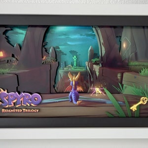 Könnte beinhalten: Gerahmtes 3D-Kunstwerk mit dem Videospiel Spyro Reignited Trilogy. Das Kunstwerk zeigt einen lila Drachen, einen goldenen Schlüssel und eine stilisierte Landschaft mit dem Logo des Spiels. Der Rahmen ist weiß.