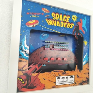Op de afbeelding: Ingelijste kunstwerk met een vintage Space Invaders arcade game scène. Het kunstwerk heeft een blauwe achtergrond met retro graphics van ruimteschepen en aliens. Het spelscherm toont gepixelde aliens en een planeet. De lijst is wit.