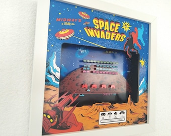 May include: Framed artwork featuring a vintage Space Invaders arcade game scene. The artwork has a blue background with retro graphics of spaceships and aliens. The game screen shows pixelated aliens and a planet. The frame is white.