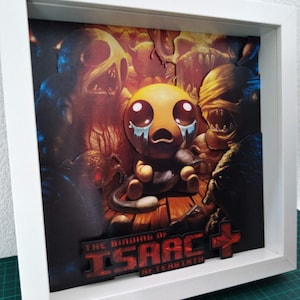 May include: A framed artwork featuring a layered paper design from "The Binding of Isaac: Afterbirth+". The artwork depicts a crying character with large eyes, surrounded by monster figures in a dark, reddish-brown background. The title is displayed in red.