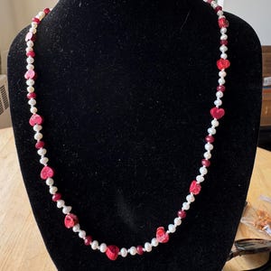 May include: A necklace with a combination of white pearls, red heart-shaped beads, and small red beads. The necklace is displayed on a black mannequin.
