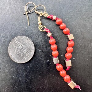 May include: A pair of dangle earrings featuring red beads and gold-colored metal accents. The earrings have a gold-colored hook and are strung on a red cord. Square gold-colored metal beads are interspersed among the red beads.