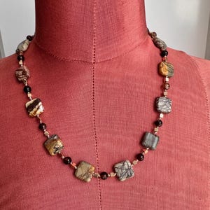 May include: A necklace featuring square and round beads in shades of brown, beige, and black. The necklace is strung on a delicate wire and displayed on a red mannequin.