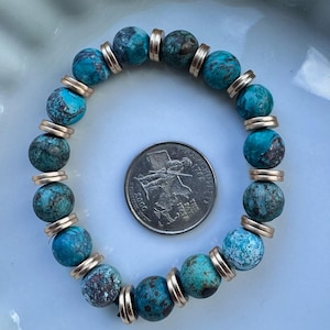 May include: A turquoise beaded bracelet with gold-colored spacers. The bracelet is made of round, textured beads in shades of blue and green. A U.S. quarter coin is next to the bracelet for size comparison.
