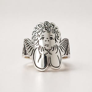 Puede incluir: Un anillo plateado con un diseño detallado de querubín. El anillo muestra la cara de un querubín con cabello rizado, manos cerca de la cara y alas a los lados. El anillo es probablemente de metal y tiene un acabado pulido.