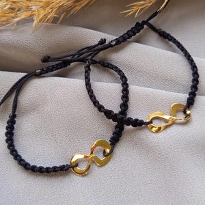 Puede incluir: Dos pulseras de macramé negro con dijes dorados con el símbolo del infinito. Las pulseras se muestran sobre una superficie gris. Las pulseras son ajustables.