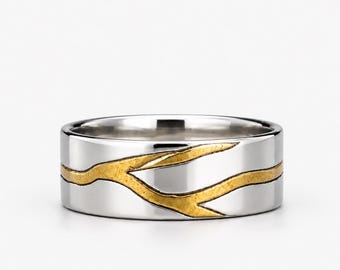 May include: A silver-toned band ring featuring a textured, gold-toned inlay that wraps around the exterior. The inlay resembles the organic, branching shape of a tree limb or twig, contrasting with the smooth, polished finish of the metal band.