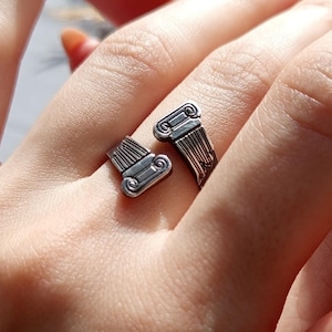 Pode incluir: Anel prateado com um design dividido, apresentando dois ornamentos em miniatura em forma de coluna. O design do anel inclui entalhes detalhados do capitel e do fuste canelado da coluna, criando uma estética grega antiga.