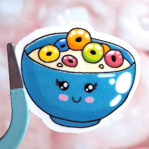 May include: A sticker featuring a cartoon bowl of cereal with a smiling face. The blue bowl is filled with milk and colorful cereal pieces. The sticker is held by a pair of tweezers against a pink background.