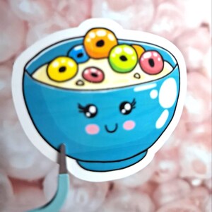 May include: A sticker featuring a cartoon bowl of cereal with a smiling face. The blue bowl is filled with milk and colorful cereal pieces. The sticker has a white border and is set against a blurred pink background.