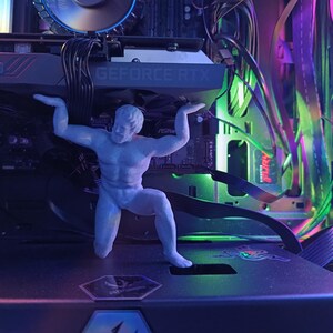 K&ouml;nnte beinhalten: Eine wei&szlig;e Plastikfigur eines Mannes in heroischer Pose, die ein Gewicht hochh&auml;lt. Die Figur steht vor einem Computergeh&auml;use mit bunten Lichtern und einer Grafikkarte mit der Aufschrift "GEFORCE RTX".