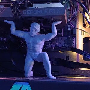 K&ouml;nnte beinhalten: Eine graue Plastikfigur einer Person, die ein Computerbauteil mit dem Text "GEFORCE RTX" hochh&auml;lt. Die Figur steht vor einem Computer-Motherboard mit verschiedenen Kabeln und Dr&auml;hten.