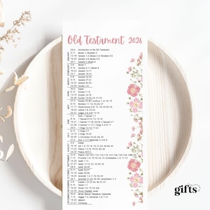 May include: A vertical bookmark with the text "Old Testament 2026" in pink. The bookmark lists Bible verses by month, with a floral border on the right side. The word "gifts" is in the bottom right corner. The bookmark is on a wooden plate.