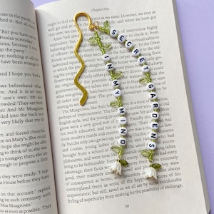 May include: A gold bookmark with green and white beads spelling out the words "Secret Garden". The bookmark is on a page of a book with text about a family gathering.