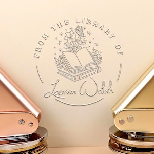Puede incluir: Dos sellos de biblioteca personalizados, uno en oro rosa y otro en oro, se muestran sobre un fondo crema. Los sellos presentan el texto "FROM THE LIBRARY OF" sobre un diseño floral y de libro, con el nombre "Lauren Walsh" debajo.